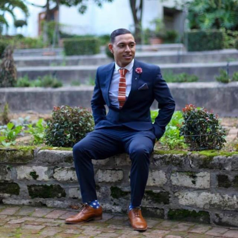 I'jaaz Van Der Schyff - Man in navy suit and striped tie sitting on stone wall in garden setting, wearing brown shoes and flower pin